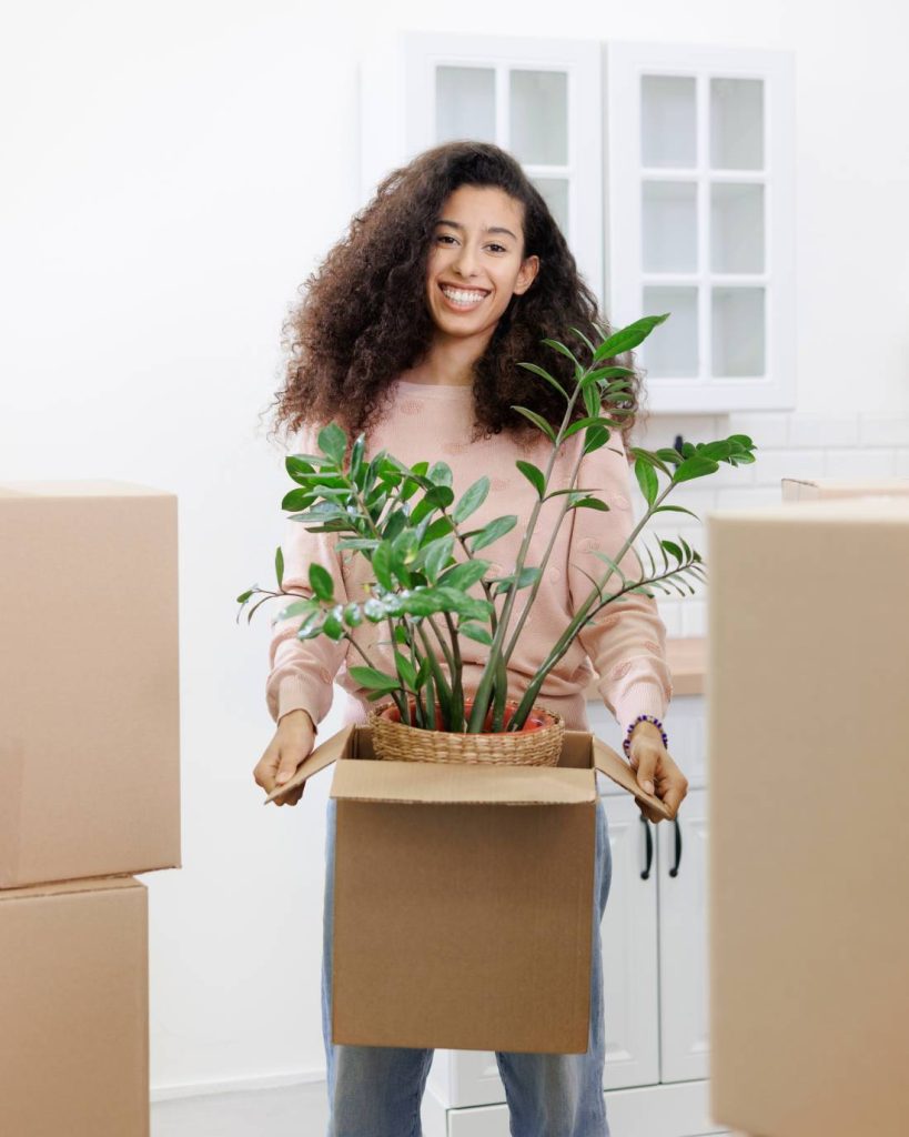 Woman with big smile hold a planted pot inside of a box. She is surrounded by moving boxes.
