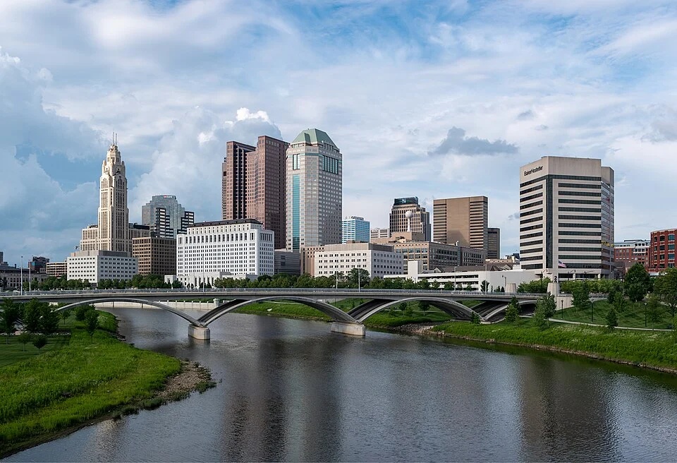 This is the downtown Columbus, Ohio skyline viewed from across the Scioto River. A multi-arched bridge spans the river in the middle, with the iconic LeVeque Tower on the left and modern office buildings behind it, all under a dramatic partly cloudy sky.